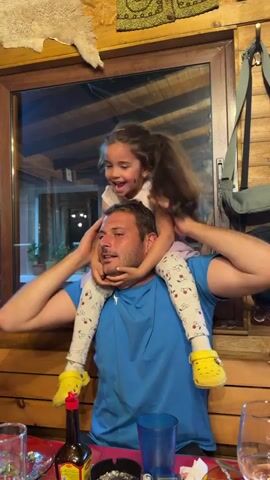A medium shot shows a father seated at a table indoors with his young daughter perched on his shoulders. The father is wearing a blue shirt, and the daughter, wearing a white top, a purple tutu, patterned leggings, and yellow Crocs, has her arms outstretched to the sides, held by someone out of frame. The father is looking directly at the camera, and the daughter is smiling broadly. A woman is seen briefly in the frame, holding a microphone up to them. The table is set with plates, glasses, a bottle of what appears to be hot sauce, and an ashtray. Behind them is a wood-paneled wall with a window showing an outdoor scene. There's also a white animal hide hanging on the wall and a patterned rug. The overall atmosphere appears festive and lively.