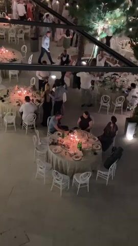 The video shows a nighttime outdoor event, likely a wedding reception, viewed from above. Several round tables are set with white tablecloths, plates, cutlery, and lit candles. People are seated at some tables and standing or dancing in the background. In the foreground, a man in a blue shirt is seated at a table, looking down and touching his face. A woman in a dark dress is seated across from him. To their left, a couple in formal attire is dancing. In the background, a group of people are dancing, some holding hands and moving in a circle. The environment appears to be a courtyard or patio with some trees and decorative lighting. The overall atmosphere seems festive and relaxed.