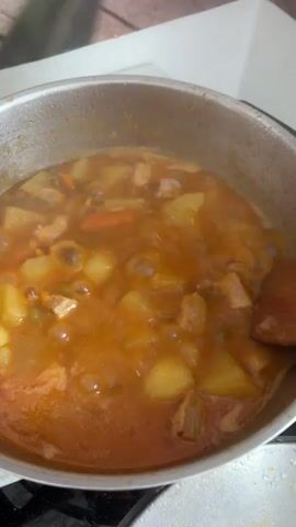 A close-up, overhead shot shows a pot of stew being stirred by a wooden spoon. The stew contains chunks of meat, potatoes, carrots, and peas in a reddish-brown broth. Steam rises from the stew, indicating it is hot. The pot is silver and appears to be made of stainless steel. The background is blurred but suggests a kitchen environment with wooden surfaces. The focus is on the food and the action of stirring.