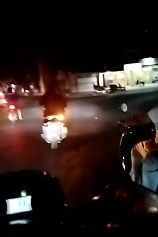 A group of people on motorcycles are riding at night. The camera is on one of the motorcycles, looking forward. The motorcycles have headlights on, illuminating the road. In the background, there are buildings and streetlights. The people on the motorcycles appear to be young. The video is shaky, suggesting the motorcycles are in motion.