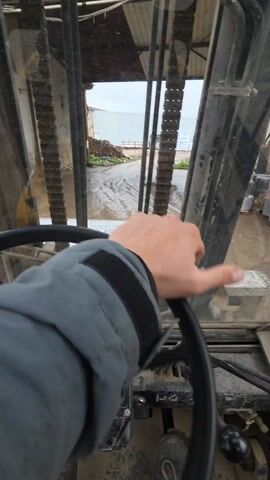 The video shows a driver's view from inside a forklift. The forklift is stationary, with the forks visible through the windshield. The steering wheel is black and dusty, with a control knob on top. The dashboard is old and has various gauges and switches. Outside, the road is muddy and wet, leading to a small building in the distance. There are stacks of materials, possibly wood or metal, on either side of the road. The weather appears overcast. A person's hand is briefly visible on the steering wheel.