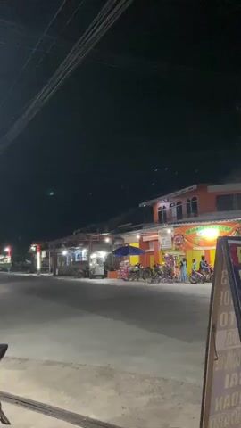 The video shows a street scene at night. There are several buildings with lights on, suggesting businesses or shops. In the foreground, there is a road and sidewalk. Several motorcycles are parked along the side of the road. There are also some people visible, appearing to be walking or standing outside. In the background, there are more buildings and streetlights. The overall atmosphere appears to be that of a small town or a suburban area at night.