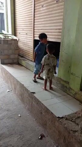 Two young boys are standing on a tiled ramp in front of a closed roll-up door. The boy on the left is wearing a blue shirt and shorts and is looking into an opening in the door. The boy on the right is wearing a patterned shirt and shorts and is also looking into the opening. The larger boy appears to be pointing or gesturing towards something inside. There is some debris on the ground around the ramp. The overall scene suggests an outdoor, possibly commercial, setting.