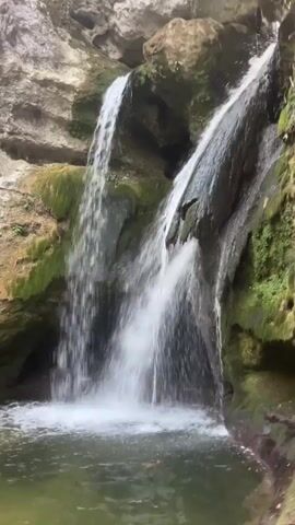 The video shows a beautiful natural scene. Two waterfalls cascade down a rocky cliff face into a clear pool of water below. The surrounding rocks are covered in moss and lush green vegetation. The water appears calm and inviting. The overall impression is one of tranquility and natural beauty.