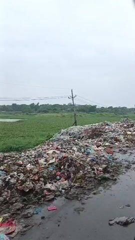 The video shows a large pile of garbage next to a wet, muddy road. In the background, there is a field of green vegetation and a cloudy sky. The garbage pile is extensive and contains a variety of waste, including plastic bags, bottles, and other debris. A single utility pole stands near the garbage pile. The camera appears to be moving, possibly from inside a vehicle, as indicated by the slight motion blur and the dark frame on the right side of the image.