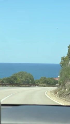 The video shows a view from inside a vehicle, looking out at a coastal road. The road curves to the right, with a guardrail on the left side. Lush green trees and vegetation line the road and the slopes above. In the distance, a body of blue water, likely the sea or ocean, stretches across the horizon. The sky is clear and blue. A few white buildings are visible nestled among the trees on the hillside. There appear to be a couple of cars on the road ahead of the camera vehicle. The overall impression is a scenic drive along a coastline on a sunny day.