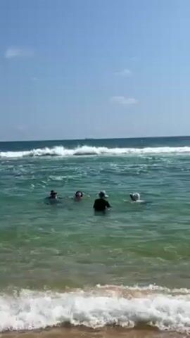 The scene shows a sunny beach with four people in the ocean. The water is a clear turquoise near the shore, gradually turning to a deeper blue further out. There are gentle waves coming in. The sky is a bright blue with a few scattered clouds. The people in the water appear to be standing or wading, as they are only partially submerged. They are spaced out from each other and seem to be enjoying the water. The beach sand is light in color. A distant ship is visible on the horizon.
