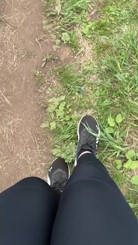 A first-person perspective shot of a person walking on a dirt path with grass on the right. The person is wearing black leggings and black and white sneakers. The camera pans down to show the person's legs and feet as they walk. The path appears to be outdoors in a natural setting.