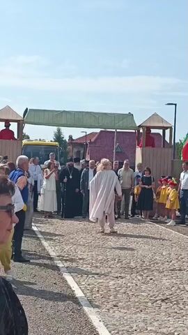 A group of people are gathered outdoors on a cobblestone surface. In the foreground, a person wearing a light-colored cloak walks away from the camera. They have long blonde hair and are wearing light-colored pants. In the middle ground, a man in a black religious robe with a pectoral cross and a staff stands among other individuals, some of whom are men in suits and others in casual attire. To the right, a group of children in yellow uniforms with red hats are visible. In the background, there are buildings with red tile roofs and a yellow bus. A wooden structure resembling a playground element with figures in red is also visible. The sky is blue and clear.