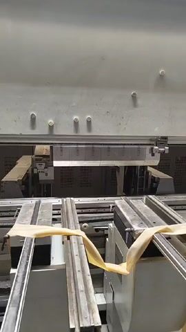 The camera faces a piece of industrial equipment in a high-angle shot. The machine is white with a metal finish and is made up of two parallel tracks with a beige strap loosely tied to the side of each track. There is a panel above with several fasteners going across. The equipment is indoors and presumably in a factory environment. The machinery is motionless throughout the video.
