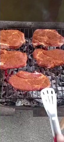 This media file shows a close-up, overhead view of six seasoned cuts of meat grilling over hot coals on an outdoor barbecue. The meat, appearing to be steaks or pork chops, is uniformly coated with a reddish-brown spice rub. As the video progresses, the meat begins to char and develop grill marks from the hot grates. A hand holding a pair of metal tongs with red handles is visible at the bottom right of the frame, occasionally used to turn or adjust the pieces of meat. The scene is set outdoors, with the grill resting on a textured surface that appears to be concrete or stone. The lighting suggests it is daytime, likely a sunny day given the brightness. There is no visible text in the video. The activity is the cooking of food on a grill, a common summertime event. The setting appears to be a residential backyard or outdoor recreational area.