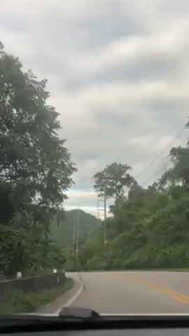 The video shows a car driving on a paved road through a hilly, forested area. The road has double yellow lines. Orange safety cones are visible on the side of the road. There are utility poles on the right side of the road. The sky is cloudy. The video is shot from the driver's perspective, and the dashboard is visible at the bottom of the frame.