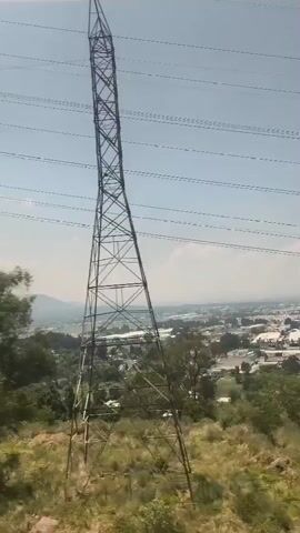 The video shows a view from a moving vehicle, passing by a metal power line tower. In the background, there's a landscape with trees, a town, and a distant mountain. The sky is clear and bright. The camera angle is from inside the vehicle, looking out the window.