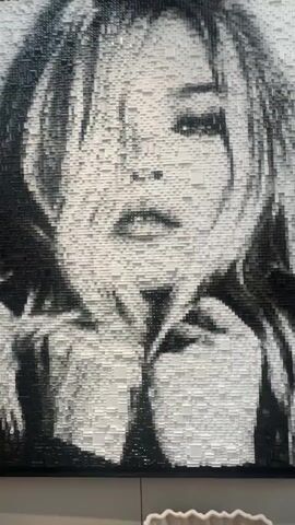 A video displays a large, monochromatic portrait of a woman's face, meticulously crafted from countless small, rectangular blocks or tiles in shades of black, white, and gray. The artwork is highly pixelated, with the varying tones and depths of the individual blocks creating the detailed image. The camera initially captures the full portrait, then zooms in to highlight the intricate construction, panning across the textured surface of the blocks. The woman depicted has flowing hair and a distinct expression, characteristic of an iconic figure. The setting appears to be an indoor art display. No discernible text is visible in the video. The location context provided is Noordwijk, Netherlands.