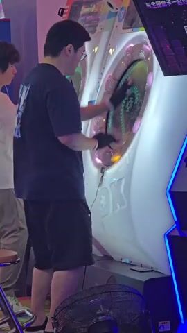 The video shows a person wearing a black t-shirt, black shorts, and black gloves playing a dance arcade game. They are standing in front of a large, white arcade cabinet with a circular screen that is displaying colorful graphics. The person's hands are on the screen, and they appear to be interacting with the game. Another person, wearing a white t-shirt and beige shorts, is standing to the left of the arcade cabinet, looking at the game. The environment appears to be an arcade, with other arcade machines visible in the background. There is also a fan on the floor in the foreground. The video captures the action from a side view.