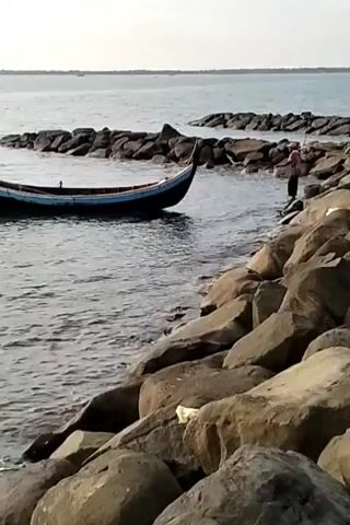The scene is at a rocky shoreline. A small, dark-colored boat with a light blue stripe is partially in the water, nestled against the rocks. The water is calm and reflects the sky, which is slightly overcast. A person is standing on the rocks near the boat, possibly interacting with it. The shoreline is made up of large, irregularly shaped rocks. In the background, the sea stretches out to the horizon, with a hint of land visible in the distance.