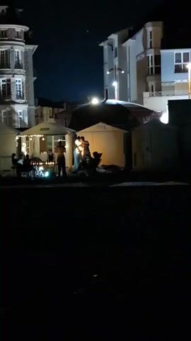 At night, silhouetted figures are gathered under a light-colored canopy structure in a dimly lit area. Several people are standing and interacting, with some appearing to be in close proximity. There are also other canopies and buildings visible in the background, illuminated by artificial lights. The ground appears to be dark and possibly sandy or pebbly. The scene suggests a social gathering or an outdoor event taking place after dark.