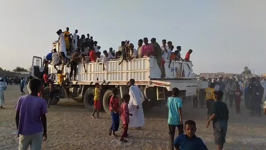Here's a description of the scene in the video:

The video shows a large group of people gathered in a dusty, open area. The focus is on a white flatbed truck that is filled with people, mostly children. Some are climbing onto the truck, while others are sitting on top or hanging onto the sides. A crowd of people, including many children, is gathered around the truck, watching or interacting with those on the truck. The people are wearing a variety of casual clothing. The sky is bright and clear, suggesting it is daytime. In the background, there are some buildings and tents, suggesting a populated area. The sound of music can be heard.