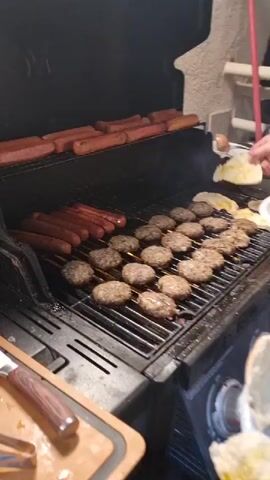 The video captures a nighttime outdoor barbecue scene in Givat Shmuel, Israel. A person is grilling a large quantity of hot dogs and hamburgers on a black gas grill. The hot dogs are arranged on two shelves, with a row of them cooking on the main grate and another row warming on the upper rack. Numerous hamburger patties are spread across the main grilling surface, sizzling and some showing char marks. A plate of what appears to be pita bread, drizzled with oil, is held by a hand in the foreground, and another plate with food is being handled near the grill. In the background, city lights are visible, suggesting an urban or suburban setting. The overall mood is casual and convivial, indicative of a social gathering or a family meal being prepared. The lighting suggests it's nighttime. No visible text is present in the video.