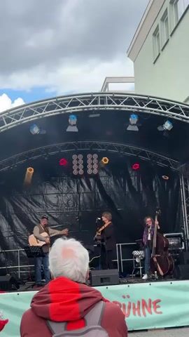 A band is performing on an outdoor stage under a black canopy illuminated by stage lights. The band consists of three musicians: one playing an acoustic guitar and singing, another playing a different acoustic guitar, and a third playing an upright bass. A person with gray hair is standing in the foreground, facing the stage, with a backpack. A sign with the word "BÜHNE" (stage) is visible below the stage. A light green building with white trim is visible in the background under a cloudy sky.