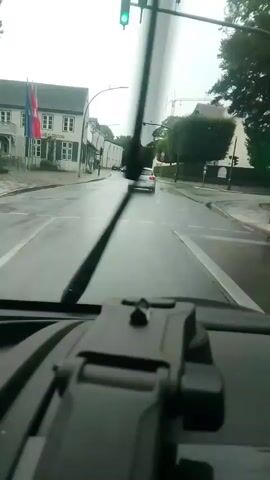 The video is filmed from inside a vehicle, looking out the windshield. Two windshield wipers are visible in the foreground, one partially obscuring the view. The road ahead is wet, indicating rain. A car is visible driving in the distance, in the same direction as the camera's vehicle. Traffic lights are visible overhead, with the green light illuminated. Buildings line the side of the road, one with flags displayed. The overall environment appears to be an urban or suburban setting during daylight hours, possibly overcast due to the wet conditions. There are no people clearly visible.