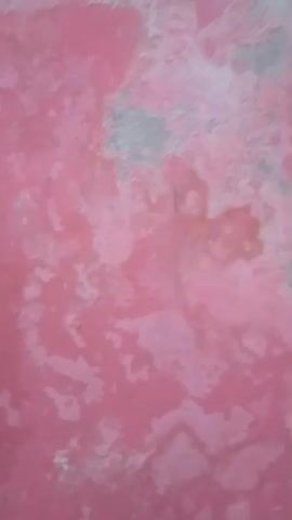 The media file displays a close-up, abstract view of a pink surface, likely a wall, with peeling paint and textured areas. The colors are predominantly shades of pink, with patches of grey and lighter pink or white where the paint has worn away. The texture is rough and uneven, suggesting age and wear. The lighting appears to be ambient, with no strong shadows indicating a specific time of day. The scene is static, with no apparent activity or subjects other than the surface itself. The location context of Gulurejo, Indonesia, does not offer any direct visual cues within the frame, as the image is an abstract textural study. There is no visible text in the media file.