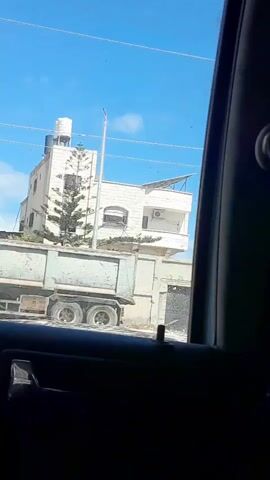 From the interior of a car in motion, the camera films a street passing by. The sky is blue with a few scattered clouds. The street is lined with a mix of residential buildings, white and gray walls, palm trees, and some overgrowth. The walls are tagged with graffiti in Arabic script. The camera is positioned in the back seat of a car and the dark frame of the car window obscures the view.