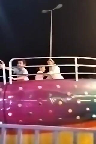 This is a short video clip of an amusement park ride at night. There are at least four people visible on the ride, which appears to be a circular car with a railing. The ride itself is brightly lit with pink and purple lights, and has white polka dots. The sky is dark. One person, a young boy in a light blue shirt, is holding onto the railing and looking forward. Another person, wearing a white shirt and head covering, is also holding onto the railing. Two other individuals are partially visible in the background. The ride is in motion, as indicated by the slight blur of the lights and the positioning of the people.