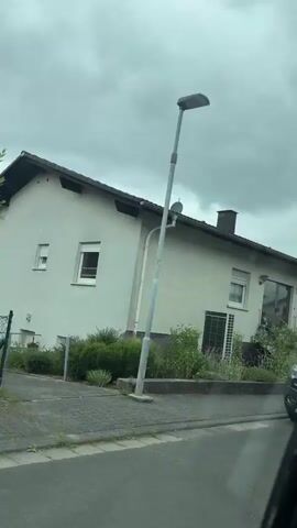 The video shows a drive-by view of houses and cars on a street. The sky is cloudy. In the foreground, a car is partially visible on the right side of the screen. The first house is light beige with a dark-tiled roof and a dark chimney. There is a garden with plants and a metal fence in front of it. A black Opel car is parked in the driveway of this house. The second house is white with a dark-tiled roof and a chimney. It has solar panels on the roof. There is a large green bush in front of the house. The third house is also white with a dark-tiled roof. It has a white rose bush in the garden. The fourth house is white with a dark-tiled roof and solar panels. A large, light-colored, flowering tree is in the garden. A black trash bin and a brown trash bin are visible next to the third house. There are steps leading up to the front doors of the houses, and some have railings. The street appears to be paved with bricks.