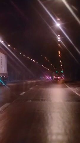 The video shows a view from inside a vehicle traveling on a wet road at night. The road is lit by overhead lights arranged in rows. There are other vehicles visible on the road ahead, including trucks and cars. The lights from the vehicles and overhead lamps create streaks and reflections on the wet surface of the road. The overall impression is of a highway or tunnel journey at night.