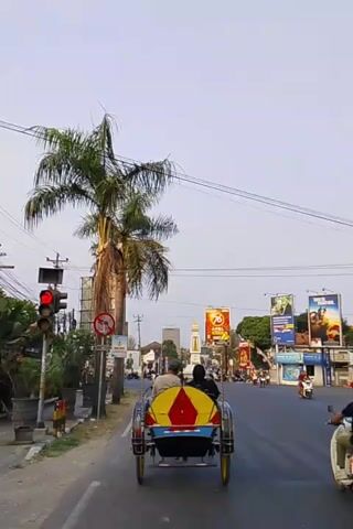 A rickshaw carrying two passengers travels down a paved road. The rickshaw is blue with yellow and red accents. Two palm trees are on the left side of the road. A traffic light is visible on the left side of the road. Billboards are on the right side of the road. The sky is clear and blue. The time of day appears to be late afternoon or early evening.