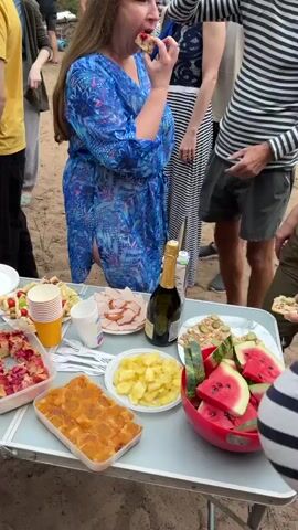 The video shows an outdoor gathering, possibly a picnic or party, with a spread of food and drinks on a long table. Several people, including men, women, and children, are present, interacting with the food and each other. The environment appears to be a sandy area with some trees and possibly a dirt road in the background.

Food items visible include various pies or cakes, sandwiches with meat and cheese, fruit platters with grapes and strawberries, sliced watermelon, and sliced cucumbers. There are also bottles of what appear to be juice, wine, and sparkling wine, along with cups and plates.

People are seen reaching for food, holding drinks, and engaging in conversation. Some individuals are dressed casually, while others are wearing more formal or themed attire, such as a person in a sailor's uniform in the last segment. The overall atmosphere seems relaxed and convivial.