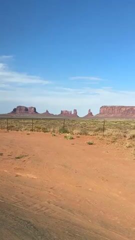 The video captures a panoramic view of Monument Valley from a vehicle's perspective. The foreground features a dusty, orange-brown dirt road with tire tracks. A wire fence stretches across the middle ground, separating the road from a vast, arid landscape. This landscape is characterized by dry, yellow-brown grasses and scattered green bushes. In the background, the iconic red rock formations of Monument Valley stand majestically against a clear, bright blue sky with a few scattered white clouds. The video appears to be shot during the daytime, suggesting fair weather. The movement of the camera indicates that the vehicle is slowly driving forward, offering a shifting perspective of the valley's monuments.