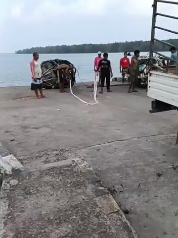 The video shows a group of men on a concrete dock near a body of water. A large, deflated, black and white object, possibly a large buoy or inflatable barrier, is on the ground, partially attached to a rope. One man is bent over, appearing to be working with the rope. Several other men stand around, observing. In the background, a truck is parked, and there is a line of trees along the water's edge. The sky is overcast. The overall scene depicts the process of moving or handling the large object.