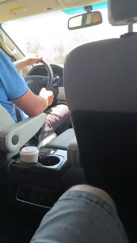 The video shows the interior of a car from the perspective of a rear passenger. The driver, wearing a blue shirt and dark pants, is visible in the driver's seat, holding the steering wheel. A cup with a lid is placed in a cup holder to the right of the driver. The background outside the car appears to be a sunny, outdoor environment with some vegetation. The video captures the driver's hands on the steering wheel and a portion of their legs.