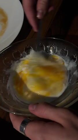 The video shows a close-up of a clear glass bowl filled with a white creamy substance and a bright yellow substance. A person's hands are holding the bowl, with one hand wearing a silver ring. Another hand is holding a dark utensil and is actively stirring the contents of the bowl, causing the yellow and white substances to swirl together. The bowl has a decorative edge with small etched heart shapes. In the background, a white plate with some residue is visible. The lighting is dim, suggesting an indoor setting, possibly a kitchen or dining area, with a wooden surface beneath the bowl. The action is focused on the preparation of the food item in the bowl, likely some sort of mixing or whisking.