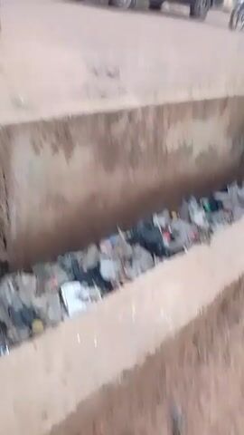 Here's a description of the video:
The video shows a concrete drainage ditch filled with trash in what appears to be a residential or commercial area. A chicken walks near the edge of the ditch. The camera pans to show a motorcycle being ridden over a makeshift bridge across the ditch. The bridge appears to be a simple concrete slab. The rider is wearing a patterned shirt and red pants.  The surroundings suggest a developing country, possibly Africa. The overall atmosphere seems ordinary and uneventful.