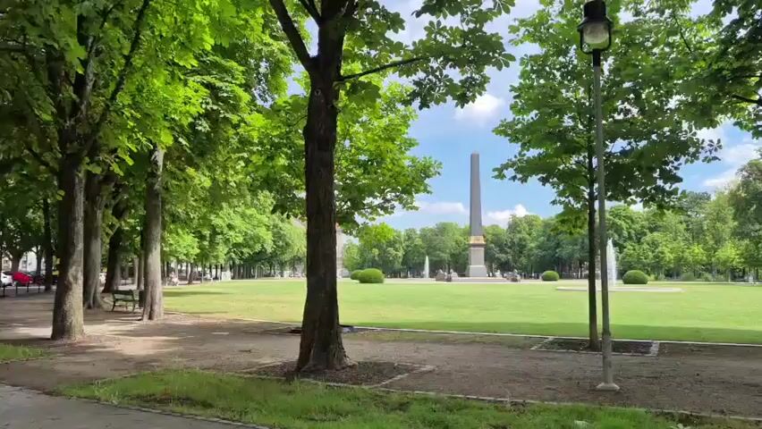 The video pans across a park with a wide green lawn and numerous trees. An obelisk stands in the background, flanked by fountains and manicured bushes. A single person sits on a park bench on the right side of the frame, facing away from the camera and appearing to read. The scene is bathed in daylight under a partly cloudy sky.
