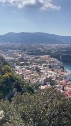 The video shows a high-angle view of a coastal town in Italy, possibly Sorrento. The town is densely packed with buildings, mostly white or light-colored, with red-tiled roofs. The town sits atop cliffs overlooking the Mediterranean Sea, which is a deep blue color. A small beach is visible along the coast. In the background, mountains are visible under a mostly clear sky with scattered clouds. The foreground is slightly out of focus, with green bushes partially obscuring the view. The camera slowly pans to the right, revealing more of the coastline.