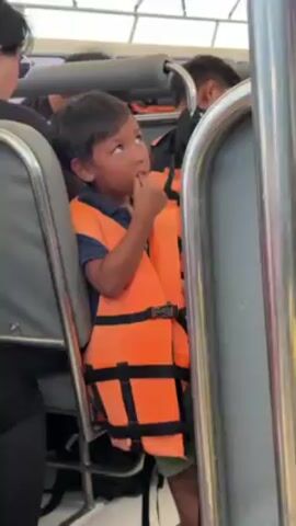 The video shows a young boy wearing an orange life vest on what appears to be a boat or ferry. He is sitting in a seat with metal framing, with other people nearby, some also wearing life vests. The boy is holding a piece of food, possibly a small piece of fruit, and seems to be looking around. The setting appears to be indoors, possibly on a partially enclosed vessel.