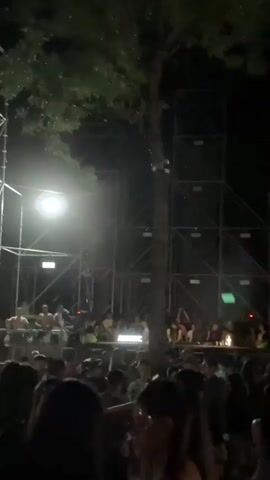 This video captures an outdoor nighttime music festival or event. A large crowd of people is gathered, facing a stage area. In the background, several tall metal truss structures are visible, supporting lighting equipment and sound systems. A prominent, tall, illuminated screen or light fixture stands to the right of the frame, displaying bright, shifting colors, primarily white and pink, with some blue appearing later. The audience is densely packed, with many individuals visible from the shoulders up in the foreground. The lighting from the stage casts an ambient glow, illuminating parts of the crowd and the surrounding trees. The overall atmosphere appears to be energetic and festive, typical of a live music performance or rave.