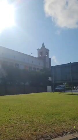 The video captures an outdoor scene with a bright sun creating lens flare. In the foreground, a paved road is visible, transitioning to a grassy area. A large billboard with blue and white text, possibly an advertisement or political campaign poster, stands prominently on the right side. The text includes "ALTERNATIV" and "PS." Further in the background, there is a building with a tower, trees, and a few vehicles, including a white van. Another billboard is visible on the left, featuring text and logos, one of which appears to say "SPP IPSP." The overall environment suggests an urban or suburban setting on a clear, sunny day.