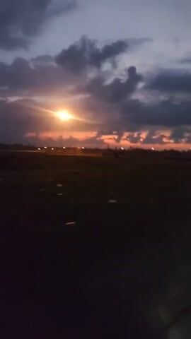 The video shows a twilight scene with a dark, open field in the foreground and a horizon with scattered lights and a line of trees or vegetation. The sky is a gradient of purple, gray, and orange, with scattered clouds. There are several streetlights or similar illuminated poles visible along the horizon. On the right side of the frame, a road with a white line is visible, with a bright light, possibly from a vehicle's headlights, illuminating the road ahead. The camera appears to be moving slightly.