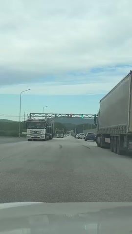 A low-angle shot from within a car on a highway. The car is stationary, and the highway stretches ahead with multiple lanes. Several large trucks are present on either side of the car's lane, with cars and other vehicles visible further ahead. An overhead gantry spans the highway, supporting traffic lights displaying both green and red signals. Distant hills and a partially cloudy sky form the backdrop. The road surface is asphalt, and the overall environment suggests a highway checkpoint or border crossing. A snippet of speech in a foreign language can be heard.