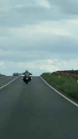 The video captures a motorcycle from behind as it travels uphill on a paved road. The rider is wearing a helmet and a jacket. Two cars are visible in the distance ahead of the motorcycle on the same road. The sky is cloudy. The right side of the road is bordered by a grassy embankment with some vegetation.