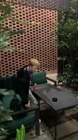 The video pans across an outdoor patio adjacent to a building constructed of red bricks. A person sits on a green velvet couch next to a small black table. The building has large glass windows displaying the interior. There is a doorway that leads into a kitchen area. Trees are on the edges of the patio.