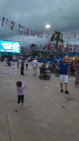 The video shows an outdoor event with a stage set up in the background. The stage is illuminated and appears to be prepared for a performance or presentation, with screens on either side. A large crowd of people is seated in chairs facing the stage, suggesting they are spectators. There are also people standing and walking around in the foreground. The environment is an open area, possibly a plaza or park, with flags strung across the top of the scene, indicating some sort of festival or international gathering. The sky is overcast and appears to be evening or late afternoon. A man in a blue polo shirt and white shorts is walking towards the camera in the foreground.