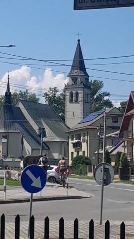 The video captures a street scene in what appears to be a European town. In the foreground, there's a dark wooden fence. A blue traffic sign with a white arrow pointing left is visible, indicating a mandatory direction. To the right of this sign, another traffic sign, possibly a warning sign, is partially obscured. The street itself is paved. On the street, a person is riding a bicycle. This person appears to be wearing shorts and possibly a backpack. Another person, wearing a backpack, is walking nearby, to the left of the cyclist. Several cars are parked or moving in the background. A light-colored car is prominent in the middle ground. Behind the street and buildings, a church with a tall, pointed steeple and a clock tower dominates the skyline. The church has a large, angled roof. Next to the church, another building with a darker roof is visible. Several houses with pitched roofs are also part of the scene, some with solar panels on their roofs. The sky is clear and blue with scattered white clouds. The overall impression is a sunny day in a populated area with architectural elements suggestive of a European setting.