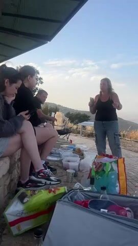 Here is a description of the video.

The video shows a group of people outdoors, standing on a brick-paved area with a view overlooking a valley. The woman is holding a can and gesturing with her left hand, apparently speaking to the others. There are coolers, bags, and containers of food and drinks scattered around the area. Some people are sitting on a low stone wall, while others are standing and listening to the woman. One man is wearing a light blue long-sleeved shirt and a white baseball cap. The sky is clear with a few clouds.