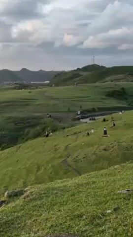 The video shows a grassy hillside landscape on what appears to be a cloudy day. Several people are sitting, standing, and walking around in different areas of the landscape. In the background, there are hills, and a tower-like structure is visible on the highest hill. There are also some motorcycles parked in the area. The overall scene depicts a recreational outdoor space with people enjoying the natural environment.